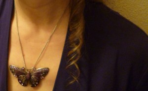 A woman's throat with a large butterfly pendant in silver and purple and a long blond ringlet on her dark garment.