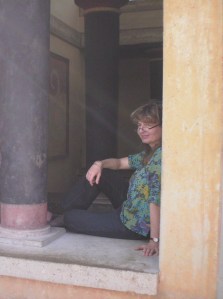 Knossos: woman in very colourful blouse sitting in a reconstructed window of the palace, in front of the same very colourful mural shown in the other photo.