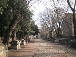 Wide avenue with sarcophagi to the left and right,leading to a mediaeval portal. Winter scene in fair weather, light-brown and light-blue.
