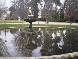 Round basin in park, with trees, houses and statue mirrored