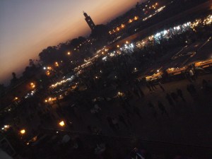 Vast square by night, illuminated by lamps in market stalls, with the sunset along the horizon and a massive minaret showing.