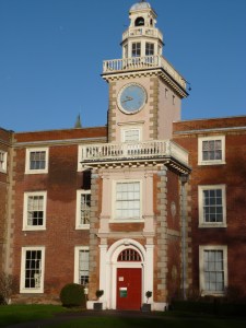Manor house in red brick, turret painted in pink, with a red door, white decorations, and a bright-blue clockface, under a blue sky.