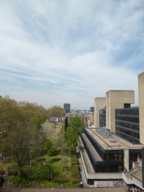 View from high up along building of concrete, steel and glass to the right, with lawn and trees to the left and clouds in blue sky above.