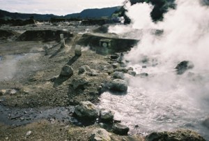 Vast surface of rough black rock to the left, gleaming pools of water and steam rising up to the right, mountains in the far background.