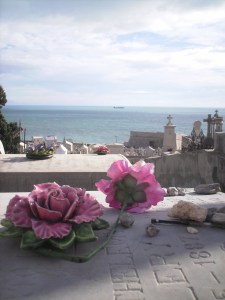 Light-blue sky and light-green ocean in the background, white tombs in the foregrund; in the front, a flat marble slabs decorated with two large pink flowers, one in clay and one in plastic.