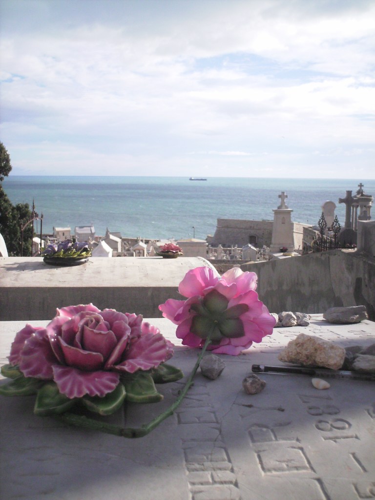 Light-blue sky and light-green ocean in the background, white tombs in the foregrund; in the front, a flat marble slabs decorated with two large pink flowers, one in clay and one in plastic.