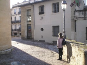Woman in winter clothes waiting under lantern in sunlit lane.